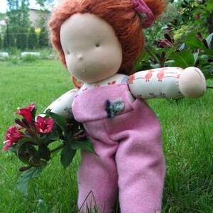 May include: A handmade doll with red hair wearing a pink overall dress and a headband. The doll is holding a bouquet of pink flowers and is standing in a grassy area.