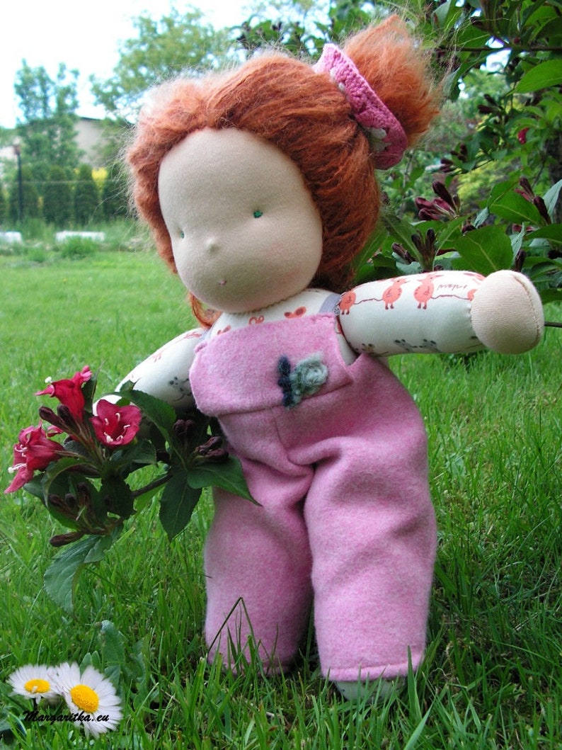 May include: A handmade doll with red hair wearing a pink overall dress and a headband. The doll is holding a bouquet of pink flowers and is standing in a grassy area.