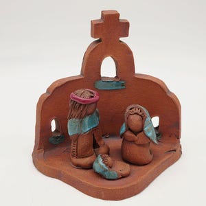 May include: A terracotta-colored ceramic nativity scene. The scene features three figures and a baby in a manger, set against a backdrop with a cross. The figures have blue and pink accents.