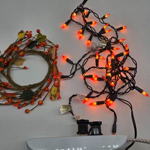 Puede incluir: Un arreglo temático de otoño con una corona decorativa con bayas y hojas naranjas, junto con una cadena de luces mini naranjas en un cable negro. Las luces están conectadas a una regleta blanca.