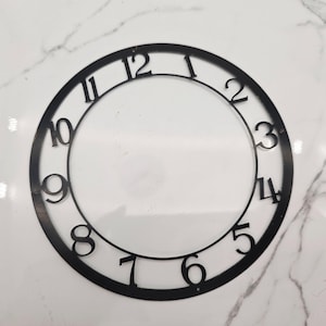 May include: A black metal clock face with the numbers 1 through 12. The numbers are in a simple, modern font. The clock face is circular with a clear center.