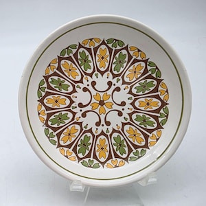 May include: White ceramic plate with a decorative floral pattern in brown, yellow, and green. The plate has a green border and a raised edge. The design is symmetrical and centered on the plate.