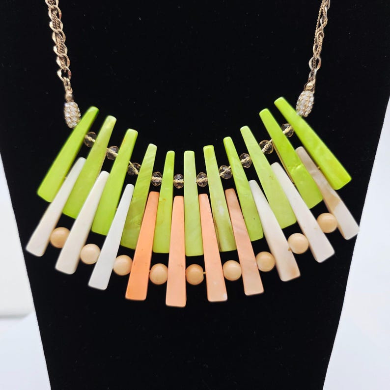 May include: A statement necklace featuring a gold chain and a pendant with a fan-like arrangement of colorful, elongated shapes. The shapes are in shades of green, white, and peach, interspersed with small, round beads. The necklace is displayed on a black surface.