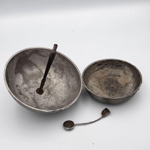 May include: Two weathered, hemispherical metal bowls with a rough, dark gray texture. One bowl features a central, upright threaded metal rod attached to its base. A separate, small, curved metal component with a flat circular end lies nearby.