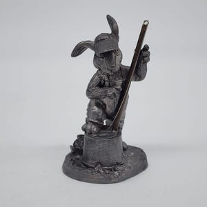 Op de afbeelding: Een gedetailleerd, zilverkleurig beeldje van een konijn met een overall en een pet, dat een lang, dun gereedschap vasthoudt. Het konijn staat op een kleine, getextureerde basis, met gedetailleerde kenmerken.