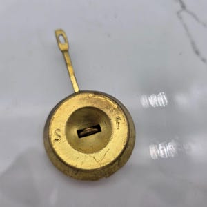 Puede incluir: Un péndulo de reloj de latón vintage. La cara circular tiene un pequeño lazo en la parte superior. La cara tiene las letras "S" y "F" a ambos lados de una abertura rectangular. El péndulo es de color dorado cálido.