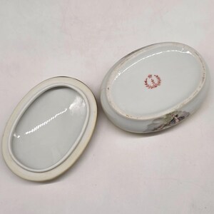 May include: An oval, white porcelain trinket box with a lid. The box features a painted scene and a red floral emblem. The lid is off, revealing the interior. The box is likely vintage.