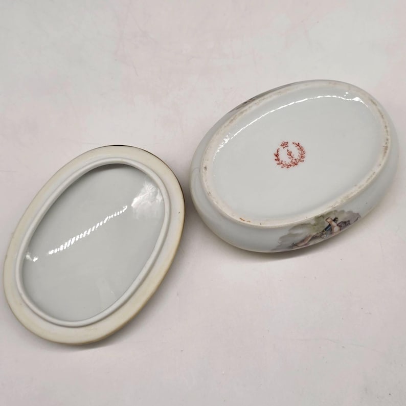 May include: An oval, white porcelain trinket box with a lid. The box features a painted scene and a red floral emblem. The lid is off, revealing the interior. The box is likely vintage.