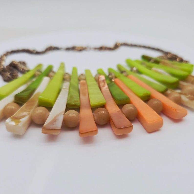 May include: A colorful necklace featuring a variety of shapes and colors. The necklace has a gold chain and is adorned with a mix of green, orange, white, and brown elements. The design includes elongated, fan-shaped pieces and round beads.