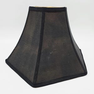 May include: A black lampshade with a square, trapezoidal shape. The shade has a ribbed texture and is trimmed with a black border. The interior appears to be a light tan color.