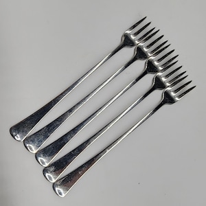 May include: Five silver forks, each with a long handle and four tines, are arranged diagonally against a white backdrop. The forks show signs of age, suggesting they are vintage or antique cutlery.