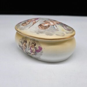 May include: Oval-shaped porcelain trinket box with a lid. The lid and body are cream-colored with gold trim. The lid features a painted scene of cherubs in a cloudy sky. The body of the box has cherub illustrations with purple accents.