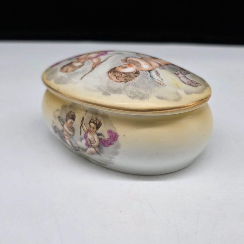 May include: Oval-shaped porcelain trinket box with a lid. The lid and body are cream-colored with gold trim. The lid features a painted scene of cherubs in a cloudy sky. The body of the box has cherub illustrations with purple accents.