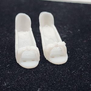 以下が含まれることがあります： 小さな白いオープントゥの靴が2足。前面にリボンがあしらわれています。靴にはわずかなヒールがあり、滑らかな素材で作られています。人形や小さなフィギュア用である可能性が高いです。