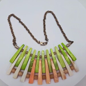 May include: A statement necklace featuring a bronze-colored chain and a pendant with green, white, and orange geometric shapes. The necklace includes small, round, light brown beads and is designed with a lobster clasp closure.