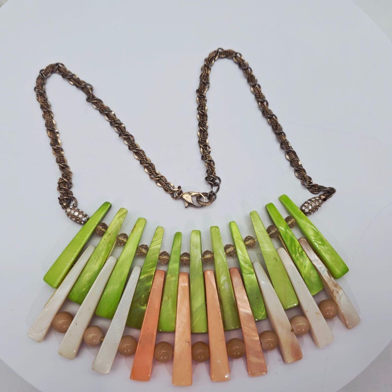 May include: A statement necklace featuring a bronze-colored chain and a pendant with green, white, and orange geometric shapes. The necklace includes small, round, light brown beads and is designed with a lobster clasp closure.