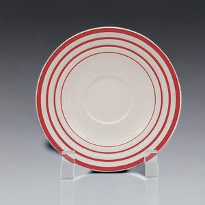 May include: A white ceramic saucer with concentric red stripes. The saucer has a central indentation and is displayed on a clear acrylic stand. The red stripes are evenly spaced and create a bold visual pattern.