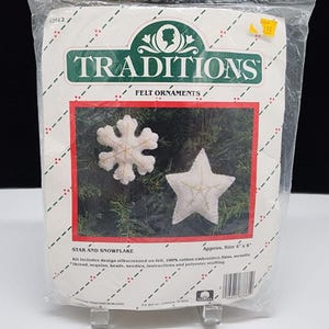 May include: A Traditions brand felt ornament craft kit in a clear plastic package. The kit includes materials to create a white snowflake and a white star ornament, each featuring gold thread detailing. The packaging text reads Star and Snowflake.