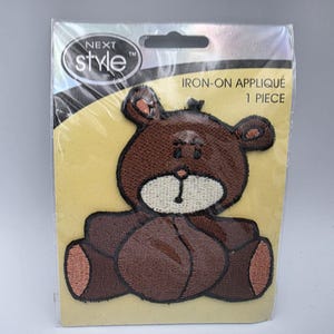 May include: A brown teddy bear iron-on appliqué in clear plastic packaging. The patch features a dark brown bear with a cream-coloured muzzle, black embroidered details, and light brown inner ears. The packaging is labelled NEXT STYLE and IRON-ON APPLIQUÉ 1 PIECE.