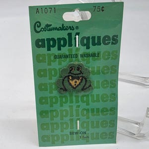 May include: A green Costumakers applique featuring a frog design with a yellow heart-shaped detail. The applique is sew-on and guaranteed washable. The packaging is green with the word "appliques" repeated multiple times.
