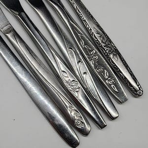 Könnte beinhalten: Eine Sammlung von Edelstahlbesteck, darunter Messer, Gabeln und Löffel. Das Besteck weist dekorative Blumen- und Blättermuster auf den Griffen auf. Die Utensilien sind diagonal vor einem weißen Hintergrund angeordnet.