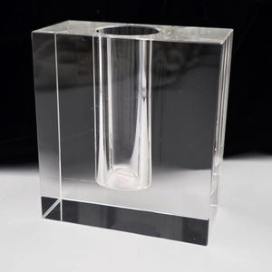May include: A clear, rectangular block vase made of solid glass. The vase features a single, cylindrical, vertical opening centered within the block, extending downward from the top surface to hold a single stem or small floral arrangement.