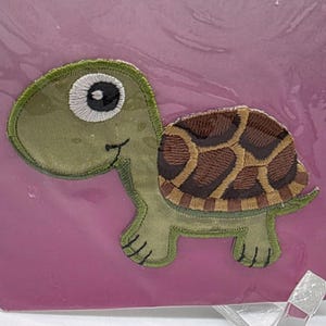 Puede incluir: Parche de tortuga bordada con cabeza y patas verdes, caparazón marrón y beige, y un ojo grande. El parche está sobre un fondo rosa.