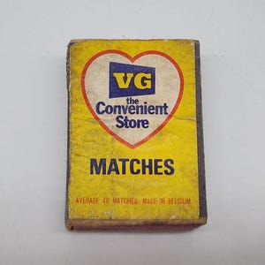 Może przedstawiać: Zabytkowe pudełko zapałek z żółtym tłem i napisem "MATCHES" w kolorze granatowym. Pudełko posiada logo w kształcie serca z napisem "VG the Convenient Store" w kolorze granatowym i białym. Widoczny jest również tekst "AVERAGE 40 MATCHES MADE IN BELGIUM".