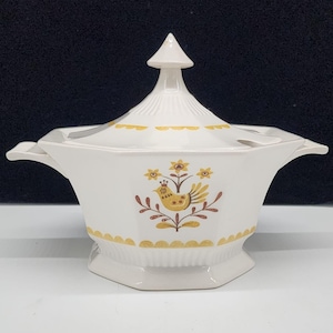 May include: White ceramic soup tureen with a lid and handles. The tureen features a yellow and brown bird and floral design. The lid has a conical finial and yellow scalloped trim. The tureen is a vintage kitchenware item.
