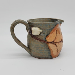 May include: Handmade ceramic pitcher with a curved handle and a spout. The pitcher is glazed in shades of blue, brown, and orange, with a leaf design on the side and a white accent near the handle. A unique piece for serving.