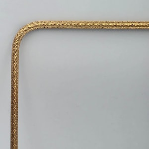 Puede incluir: Un marco rectangular de color dorado que presenta una esquina redondeada. El marco tiene una superficie texturizada y en relieve con un patrón repetitivo de pequeñas líneas diagonales y puntos a lo largo de toda su longitud.
