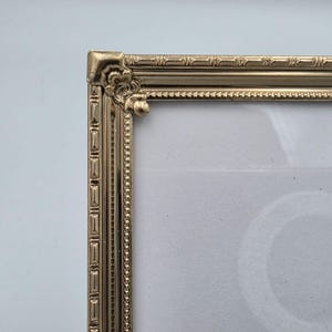 May include: A gold-toned metal picture frame corner featuring an ornate, raised scrollwork detail at the joint. The frame edge displays a repeating rectangular pattern alongside a delicate beaded border, surrounding a plain white insert.