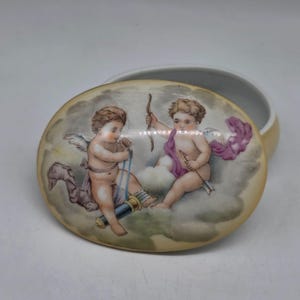 May include: A small, oval-shaped trinket box with a lid featuring a painted scene of two cherubs with bows and arrows. The cherubs are depicted in a cloud-filled sky, with soft pastel colors and delicate details. The box has a cream-colored base.