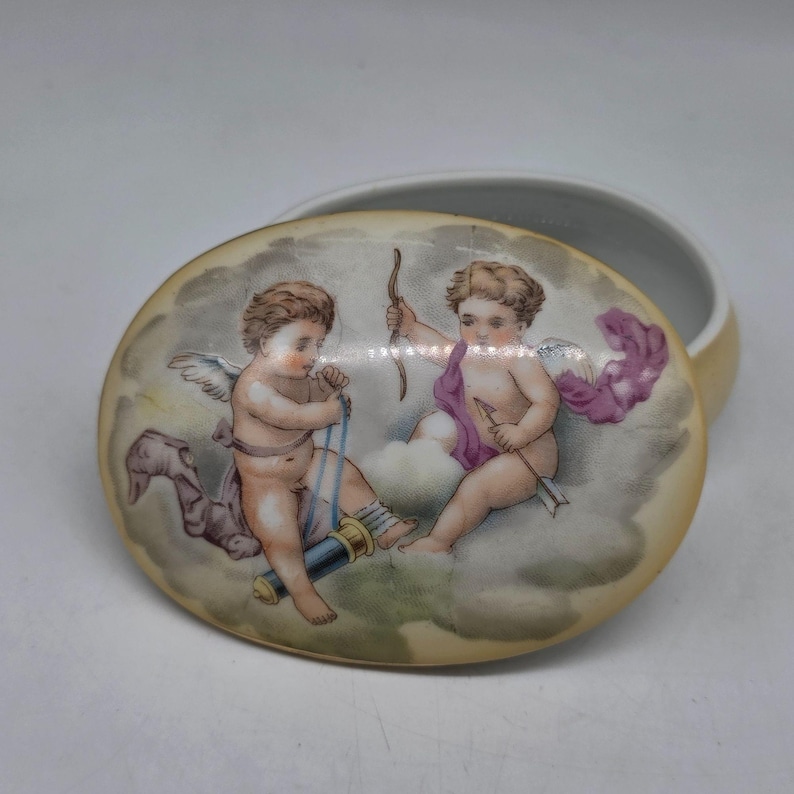 May include: A small, oval-shaped trinket box with a lid featuring a painted scene of two cherubs with bows and arrows. The cherubs are depicted in a cloud-filled sky, with soft pastel colors and delicate details. The box has a cream-colored base.