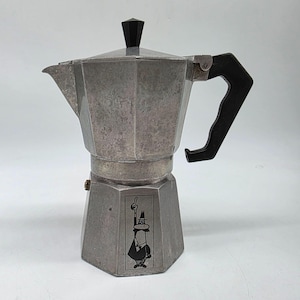May include: A silver stovetop espresso maker with a black handle and lid knob. The coffee pot has a geometric design and a black and white graphic on the base.