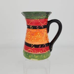 May include: A colorful ceramic pitcher with a black handle. The pitcher features horizontal bands of red, orange, and black, with decorative yellow accents. The top and bottom rims are green. The pitcher is on a clear stand.
