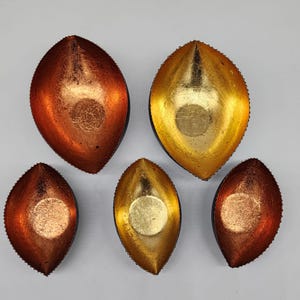 May include: Five decorative leaf-shaped bowls in shades of gold and copper. The bowls have a textured metallic finish and a small, circular indentation in the center. The edges are lined with small, decorative beads.
