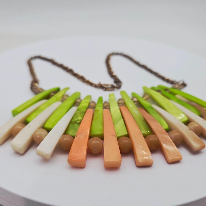 May include: A statement necklace featuring a bronze chain and a fan-shaped design. The necklace showcases a vibrant array of colors, including white, light green, and orange, with small, round, tan beads.