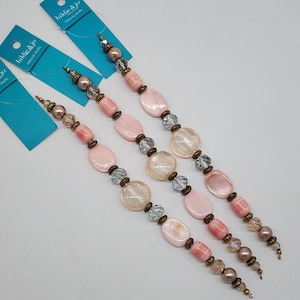 May include: Three strands of natural shell beads in shades of pink, cream, and brown. The beads are strung together with small metal accents and clear crystal beads. Each strand has a blue tag that reads "hildie & jo natural shells."