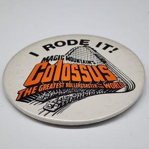 May include: A round, off-white button with the text "I RODE IT!" above a graphic of a roller coaster. The button features the words "MAGIC MOUNTAIN'S COLOSSUS THE GREATEST ROLLERCOASTER IN THE WORLD" in orange and black.