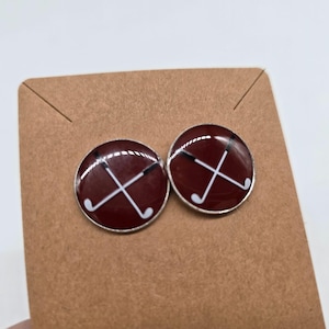 Può includere: Un paio di orecchini a bottone rotondi a tema golf. Ogni orecchino presenta uno sfondo rosso scuro con un design di mazze da golf bianche incrociate e un bordo argentato. Gli orecchini sono esposti su una carta marrone.
