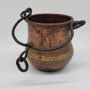 Może przedstawiać: Mały, antyczny kociołek miedziany z ciemnym, kutym żelaznym uchwytem. Kociołek ma zaokrąglony korpus z pasem wyblakłej złotej farby pośrodku. Miedź ma bogatą, postarzaną patynę.