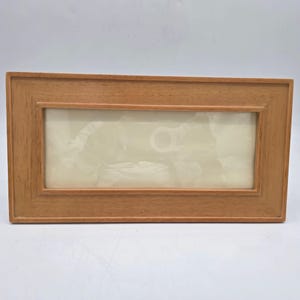 May include: A rectangular wooden picture frame with a light brown finish. The frame has a simple, classic design with a rectangular opening for a photograph or artwork. The frame is designed to be displayed on a wall or tabletop.
