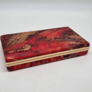 May include: Rectangular box with a red, brown, and tan abstract pattern. The box has a gold-colored trim around the edges. The box is closed and sits on a white surface.