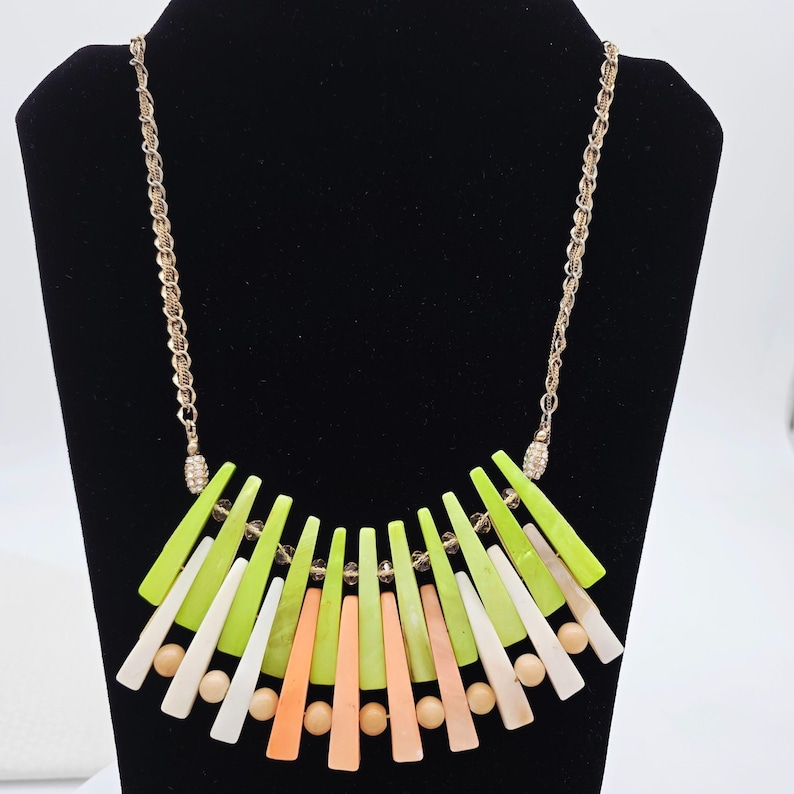 May include: A statement necklace with a gold-tone chain and a pendant featuring a fan-like arrangement of colorful geometric shapes. The shapes are in shades of green, white, peach, and beige, with small round beads and clear crystal accents.