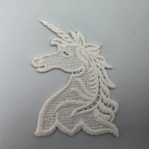 May include: A white embroidered unicorn head patch featuring a detailed horn, a flowing mane, and a stitched profile silhouette. The design is crafted with dense thread work, creating a textured surface that highlights the mythical creature's form.