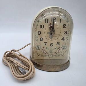 May include: Vintage GE electric alarm clock with a cream-colored face and a clear, domed cover. The clock face features black numerals, floral accents, and the words "General Electric". A tan power cord extends from the clock.