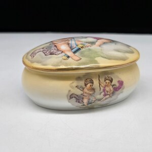 May include: Oval-shaped ceramic trinket box with a lid. The box is a soft yellow with a gold trim. The lid and body feature painted cherubs and clouds in pastel colors. A decorative item for storing small items.