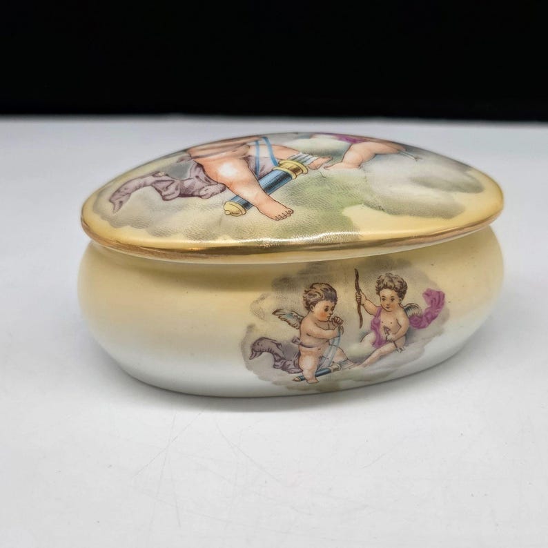 May include: Oval-shaped ceramic trinket box with a lid. The box is a soft yellow with a gold trim. The lid and body feature painted cherubs and clouds in pastel colors. A decorative item for storing small items.