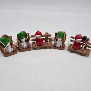 May include: Five miniature wooden Christmas ornaments. Each features a small figure in a red or white outfit and a matching hat, seated on a wooden bench or swing. The ornaments are strung with twine, ideal for festive decoration.